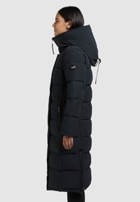 Abrigo acolchado largo en azul marino oscuro con diseño acolchado, cuello alto y capucha ajustable. Presenta cremalleras laterales y un logo en la manga.