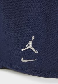 Marineblå stofkasket med sølvbroderede Jumpman- og Nike-logoer. Materialet har en tekstureret overflade med en struktureret form.