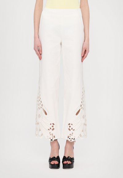 Pantalons larges blancs avec des motifs découpés détaillés aux ourlets, présentant une texture lisse et de subtils accents marron. Associés à des chaussures à plateforme noires.