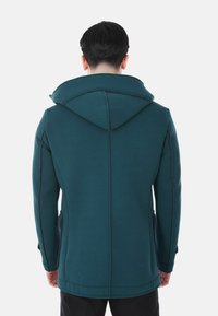 Uomo che indossa una giacca con cappuccio verde acqua scuro con dettagli di cucitura visibili, in piedi rivolto di spalle su uno sfondo bianco.