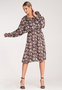 Czarna kwiecista sukienka z długimi rękawami, marszczoną talią i długością do kolan. Zawiera różowe i białe wzory kwiatowe, noszona z czarnymi sandałami na obcasie.