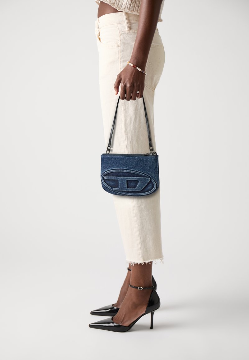 Borsa in denim con un logo ovale prominente, dotata di chiusura a zip e manici sottili, abbinata a scarpe nere con tacco alto e pantaloni di colore chiaro.