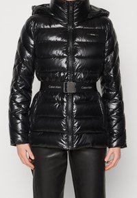 Chaqueta acolchada negra brillante con capucha, cremallera frontal, bolsillos laterales y un cinturón de marca Calvin Klein alrededor de la cintura, combinada con pantalones negros.
