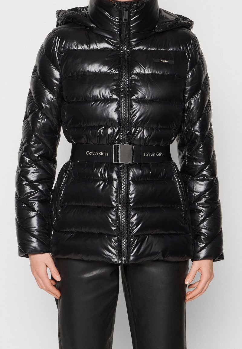 Chaqueta acolchada negra brillante con capucha, cremallera frontal, bolsillos laterales y un cinturón de marca Calvin Klein alrededor de la cintura, combinada con pantalones negros.