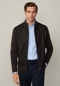 Veste zippée marron foncé à manches longues, texture côtelée et col montant, portée par-dessus une chemise bleu clair à motif à pois.
