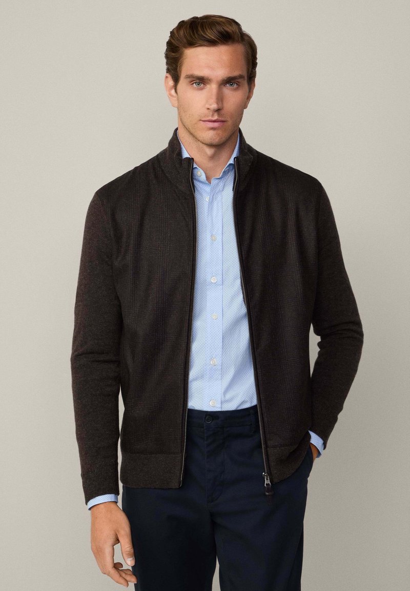 Veste zippée marron foncé à manches longues, texture côtelée et col montant, portée par-dessus une chemise bleu clair à motif à pois.