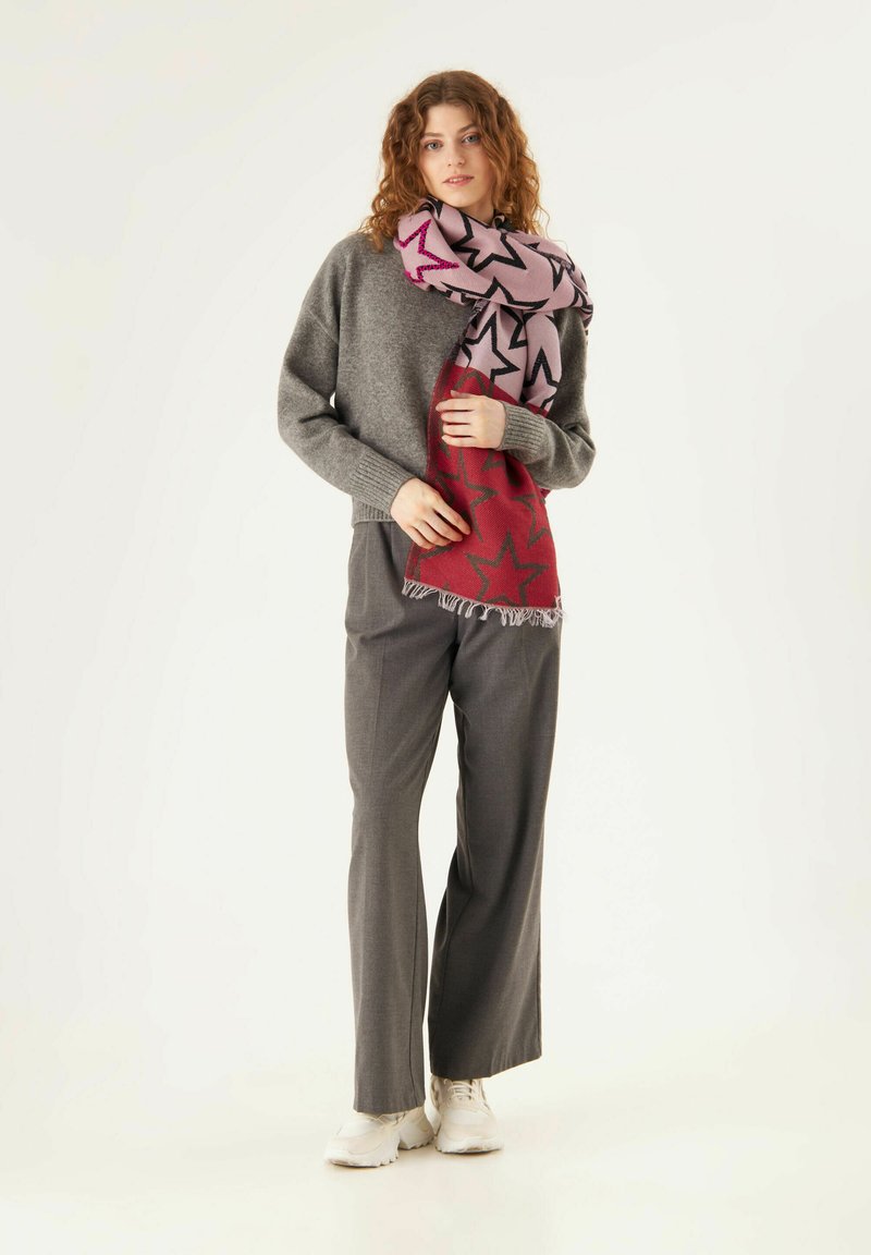 Pull en tricot gris, pantalon large gris, et une écharpe à motifs étoiles rose-rouge avec des franges. Le mannequin porte des baskets blanches.
