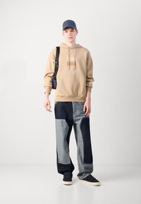 Felpa beige con cappuccio e logo, cappellino blu e ampi pantaloni di jeans a pezzi nei toni chiari e scuri. Le sneakers completano l'outfit.