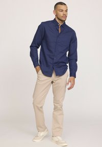 Chemise à manches longues marine avec col boutonné et poche poitrine, associée à un pantalon beige clair et des baskets blanches, dans une pose debout.