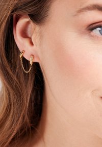 Boucles d'oreilles en or en forme de hoop avec une chaîne reliant deux pièces. Le premier anneau est lisse, tandis que le second comporte de petits cristaux incrustés.