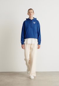 Felpa blu con cappuccio e coulisse, logo bianco, indossata sopra una maglietta bianca. Abbinata a pantaloni beige larghi e scarpe da ginnastica bianche. Sfondo semplice.