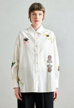 Chemise blanche en coton ornée d'une broderie florale colorée sur les manches et la poitrine, col classique, devant à boutons, coupe longue et ample.