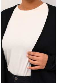 Sort cardigan over en hvid ribbet top, der har en V-udskæring og lange ærmer. Cardiganens stof ser blødt og letvægtsagtigt ud.