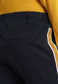 Pantalon marine en tissu texturé, doté d'une coupe slim, d'une taille élastique et de bandes latérales blanches et moutarde. Une poche arrière visible.