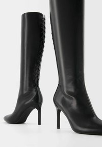 Knähöga svarta läderstövlar med spetsig tå, hög stilettklack och snörningsdetaljer längs baksidan för extra textur.