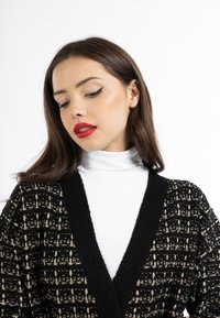 Cardigan nero e oro con texture, con scollo a V profondo e maniche lunghe, indossato sopra un dolce collo alto bianco. Materiale liscio e lavorato a maglia.