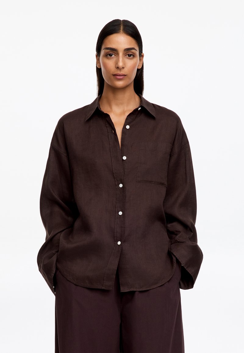 Femme aux cheveux foncés porte une chemise marron foncé ample boutonnée et un pantalon assorti, debout les mains dans les poches sur un fond blanc.