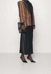 Blazer marron à motif serpent avec revers noirs, associé à une longue jupe en satin noire et des talons pointus noirs avec des accents vert néon.