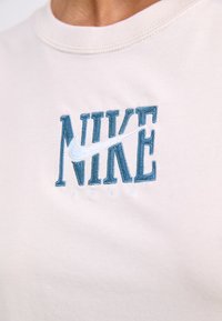 T-shirt en coton beige arborant un logo "NIKE" brodé en bleu avec un swoosh blanc et l'année "1972" en dessous.
