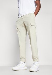 Pantalon cargo beige avec des chevilles élastiques, deux poches latérales et une taille avec cordon de serrage, associé à des chaussures de sport blanches.