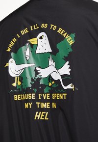 Chemise noire avec texte jaune autour d'une carte verte, présentant trois mouettes blanches, dont une tenant un poisson, avec les mots « Quand je mourrai, j'irai au paradis parce que j'ai passé mon temps en enfer. »