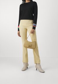 Osoba nosząca czarny sweter z kolorowymi mankietami, żółte teksturowane spodnie, beżowe botki za kostkę i trzymająca pasującą żółtą torebkę.