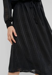 La main d’une femme repose à côté d’une jupe noire transparente avec des rayures verticales et diagonales, attachée à la taille avec un cordon.