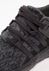 Svart sportig sko med texturerad stickad ovandel, svarta snören och plastdetaljer. Designen inkluderar subtila tonala mönster och ett lager-på-lager-utseende.