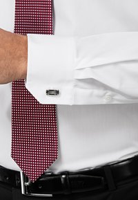 Witte overhemd met Franse manchetten, zilveren manchetknoop en een kastanjebruine stropdas met een klein vierkant patroon. Zwarte leren riem zichtbaar.