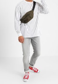 Camicia bianca a maniche lunghe con righe nere, jeans slim-fit grigi e sneakers rosse. Borsa verde a tracolla con dettaglio della zip frontale.