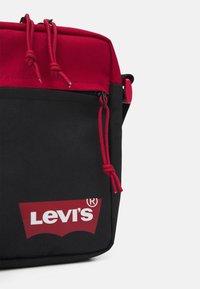 Svart och röd Levi's axelväska med en strukturerad design, som har en röd övre del, dragsnören och en logotyp på framsidan.