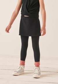 Schwarzer Rock über schwarzen Leggings, strukturierte graue Sneakers mit weißen Sohlen und korallenfarbene gerippte Bündchen, gestylt mit einem verdrehten schwarzen Oberteil.