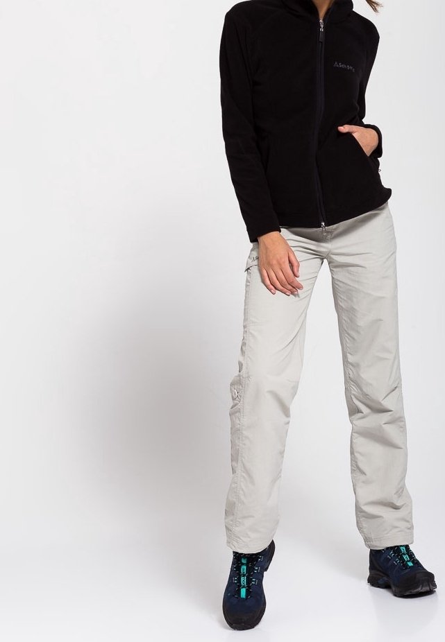 Femme portant une veste noire zippée, un pantalon cargo gris clair et des chaussures de randonnée foncées, debout devant un fond uni.