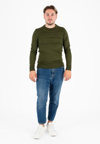 Man staat, draagt een olijfgroene lange mouwen shirt, blauwe spijkerbroek en witte sneakers, tegen een effen witte achtergrond.