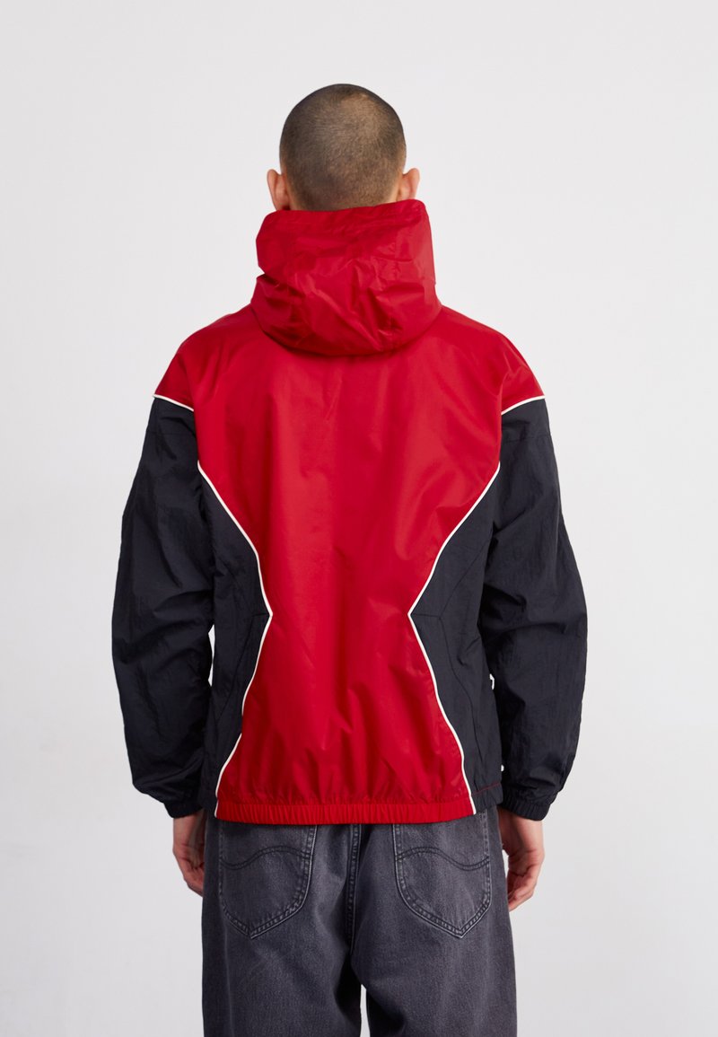 Giacca a vento rossa e nera con collo zip, polsini elastici e cappuccio con cordino. Il design include cuciture bianche a contrasto.