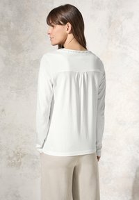 Weißes Langarmshirt mit glatter Textur, ausgestattet mit einer Pleat hinten und dekorativen Nieten entlang des Ausschnitts, kombiniert mit beigen Hosen.
