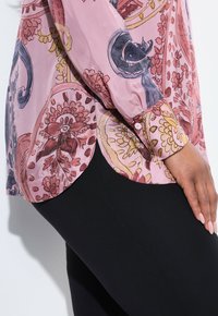 Transparente, rosa Bluse mit floralen Mustern in Rot, Gelb und Blau, mit abgerundetem Saum und geknöpften Manschetten, kombiniert mit schwarzen Leggings.