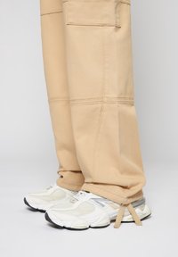 Beige cargobyxor med synliga sömnadsdetaljer, parat med vita och grå sneakers med mesh- och gummidetaljer.