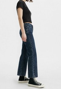 Jean en denim foncé à coupe large et taille haute, associé à un t-shirt noir court et des baskets montantes noires. Design épuré et décontracté.