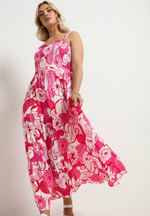 Femme portant une robe maxi fluide sans manches à fleurs roses et blanches, tenant un tissu, debout sur un fond clair uni.