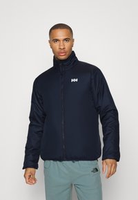 Chaqueta acolchada de color azul marino con un cuello alto y cremallera frontal completa. Presenta un logo blanco en el lado izquierdo del pecho y puños elásticos para un ajuste ceñido.