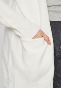 Bílý cardigan s ribbed manžetami a lemem, s bočnými kapsami. Materiál vypadá měkký s texturovaným povrchem.