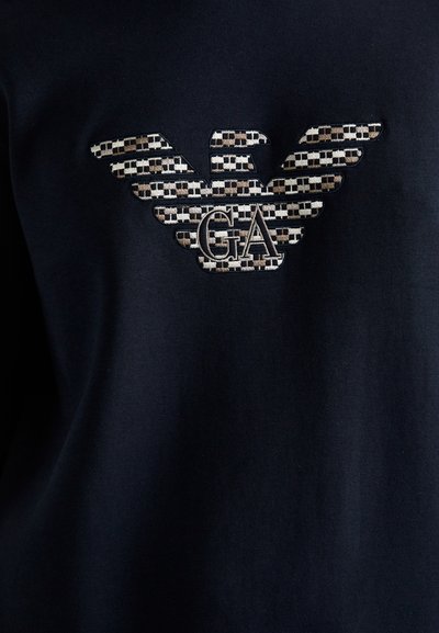 Zwarte stof met geborduurd geruit adelaar logo en de letters "GA" in het midden, dat de Giorgio Armani branding vertegenwoordigt.