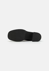Suela de zapato negra con una superficie de goma texturizada. El tacón es grueso y se eleva ligeramente en la parte posterior. Parte delantera plana con un acabado suave.