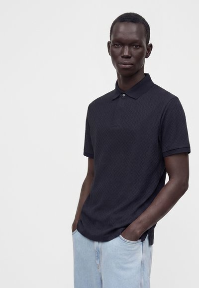 Homme portant un polo à manches courtes texturé sombre et un jean bleu clair, debout les mains dans les poches devant un fond uni.