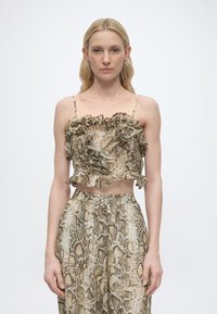 Γυναίκα με ταιριαστό τοπ crop με βολάν και παντελόνι ψηλόμεσο, και τα δύο με τύπωμα φιδιού, στέκεται μπροστά σε απλό φωτεινό φόντο.