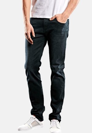 Man wearing black slim-fit jeans, white sneakers, and a white shirt, standing with one hand in pocket against plain background.