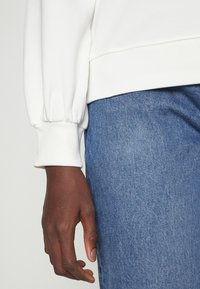 Felpa bianca con polsini a costine e manica leggermente gonfia, abbinata a jeans di denim blu con trama.