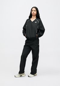 Fekete Nike melegítőfelső V-nyakú pulóverrel és elasztikus derékpántos nadrággal. Fehér csíkozással és logóval rendelkezik. Az anyag könnyűnek és simának tűnik.