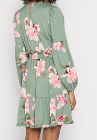 Robe fleurie en tissu vert clair, taille élastique, manches longues bouffantes, ourlet à volants et ouverture dos en forme de goutte.